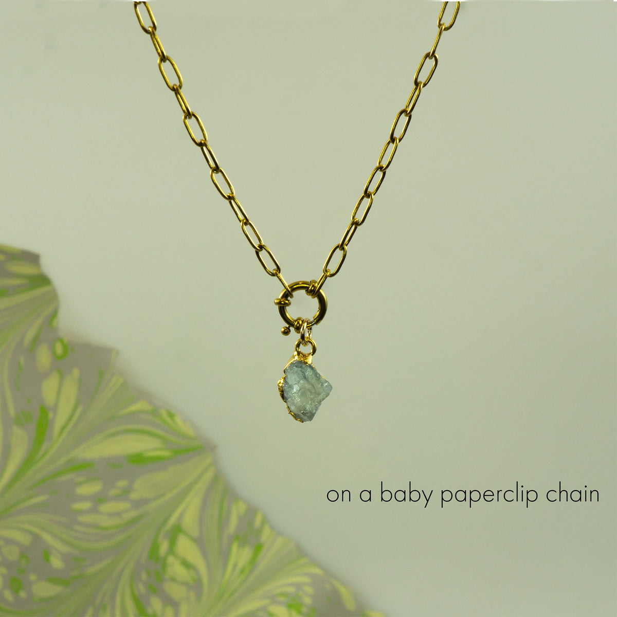 A gold paperclip chain necklace with a small gold clasp holding a raw, irregular light blue aquamarine gemstone pendant