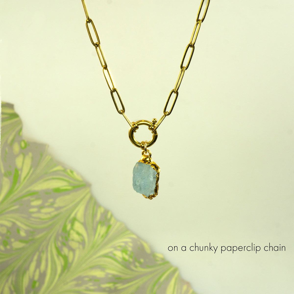 A gold paperclip chain necklace with a gold ring clasp holding a chunky, raw light blue aquamarine stone pendant