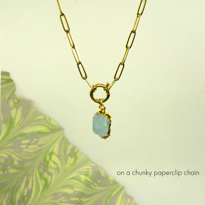 A gold paperclip chain necklace with a gold ring clasp holding a chunky, raw light blue aquamarine stone pendant