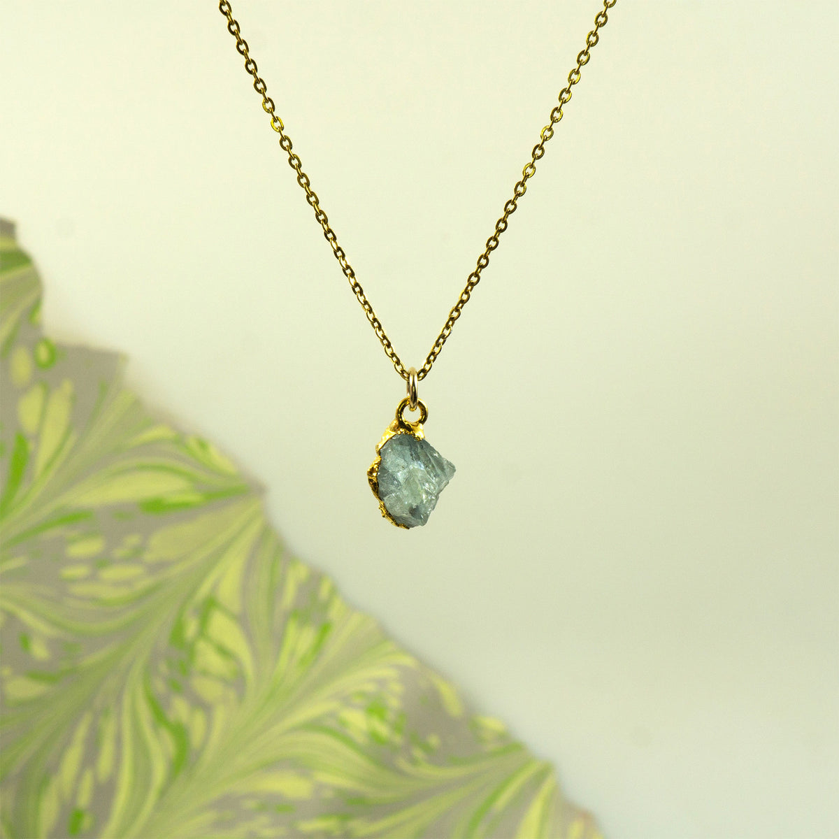 A gold chain necklace with a raw, irregular light blue aquamarine gemstone pendant