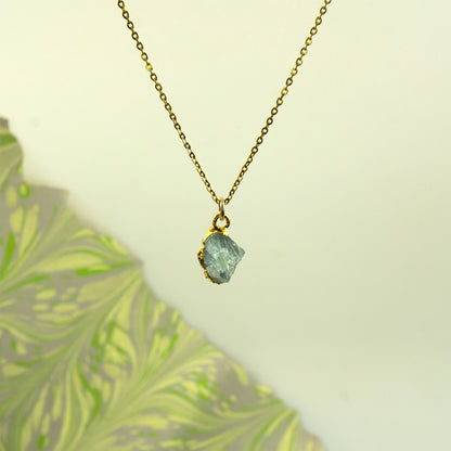 A gold chain necklace with a raw, irregular light blue aquamarine gemstone pendant