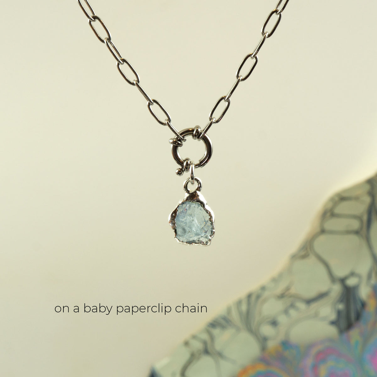 A silver paperclip chain necklace with a small ring clasp holding a rough, irregular aquamarine gemstone pendant