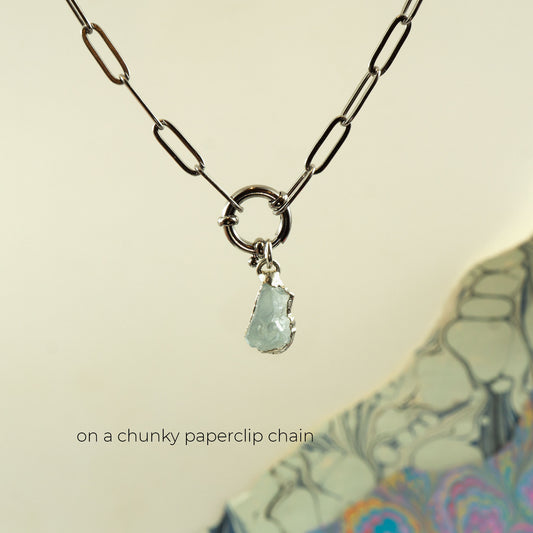 A silver paperclip chain necklace with a circular ring connector and a small raw, irregular-cut pale aquamarine gemstone pendant