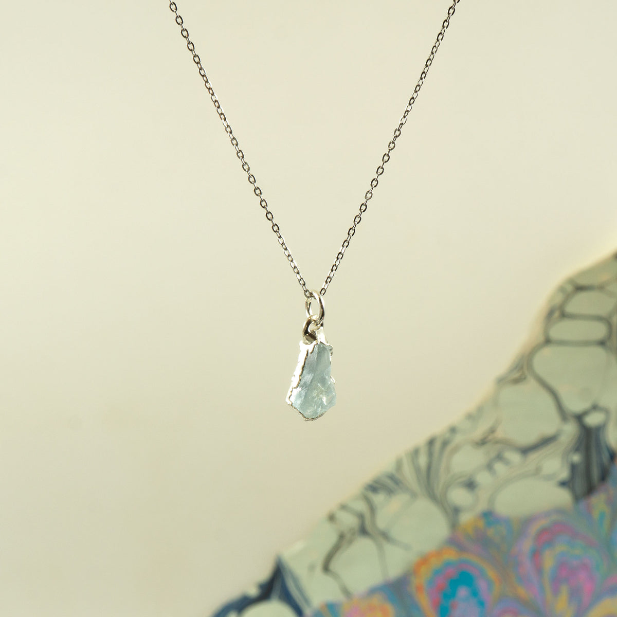 A delicate silver chain necklace with a small, irregular faceted light blue aquamarine gemstone pendant