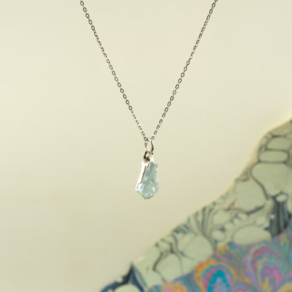 A delicate silver chain necklace with a small, irregular faceted light blue aquamarine gemstone pendant