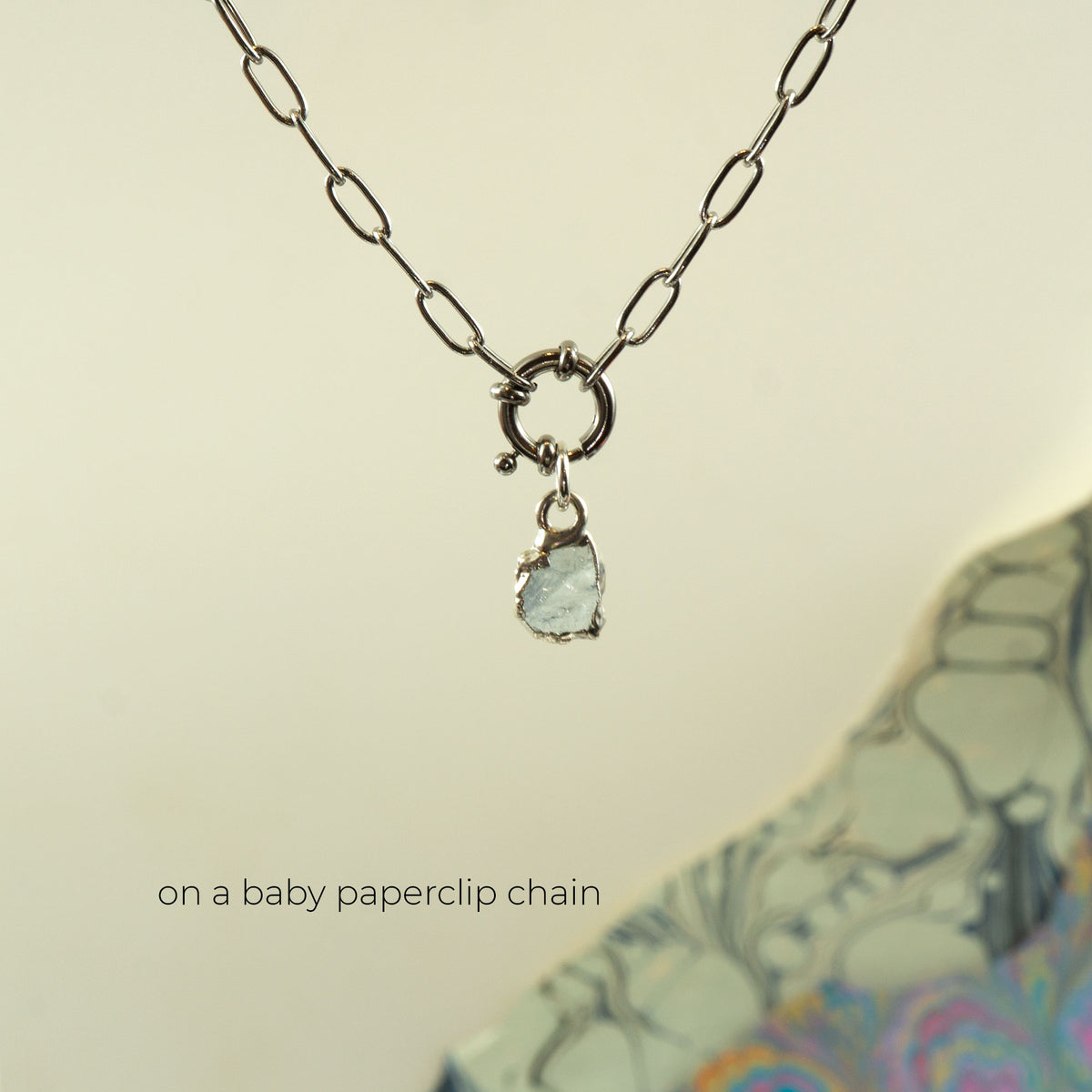 A silver paperclip chain necklace with a small ring clasp and a dangling light blue aquamarine gemstone charm