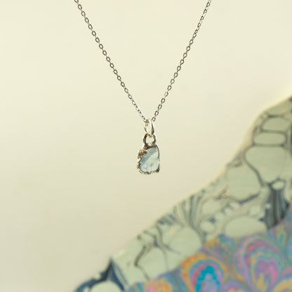 A delicate silver chain necklace with a small, irregular-cut pale blue-green aquamarine stone pendant.