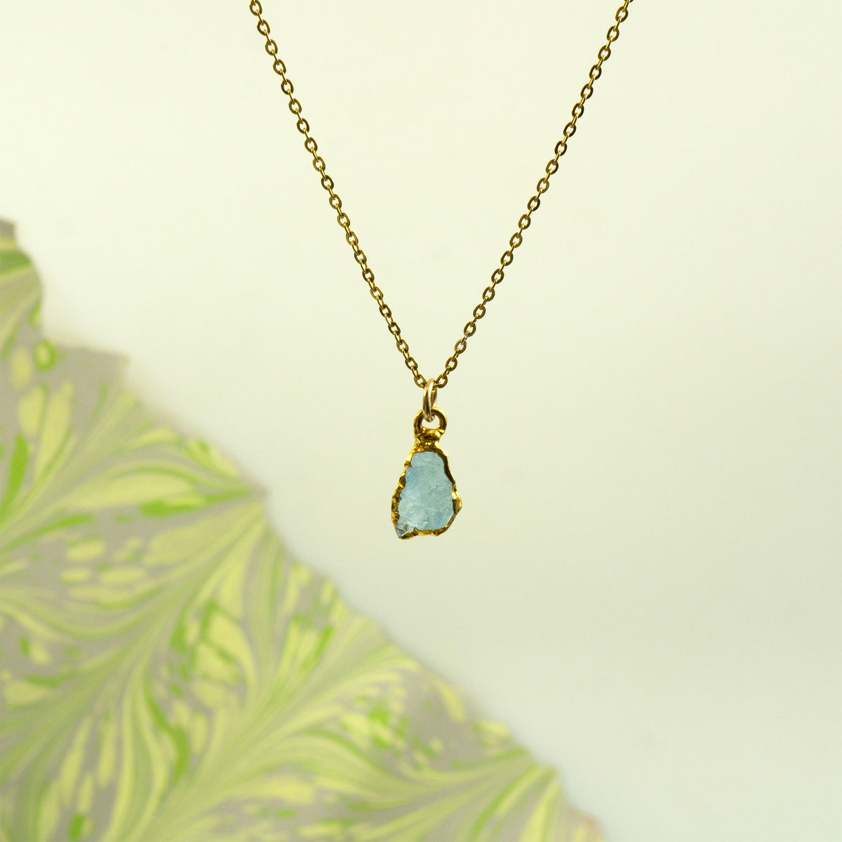 A gold chain necklace with a raw, irregular light blue aquamarine gemstone pendant