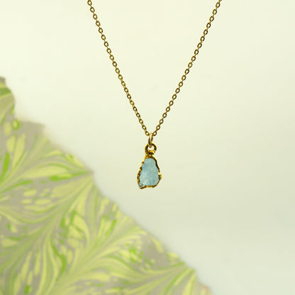 A gold chain necklace with a raw, irregular light blue aquamarine gemstone pendant