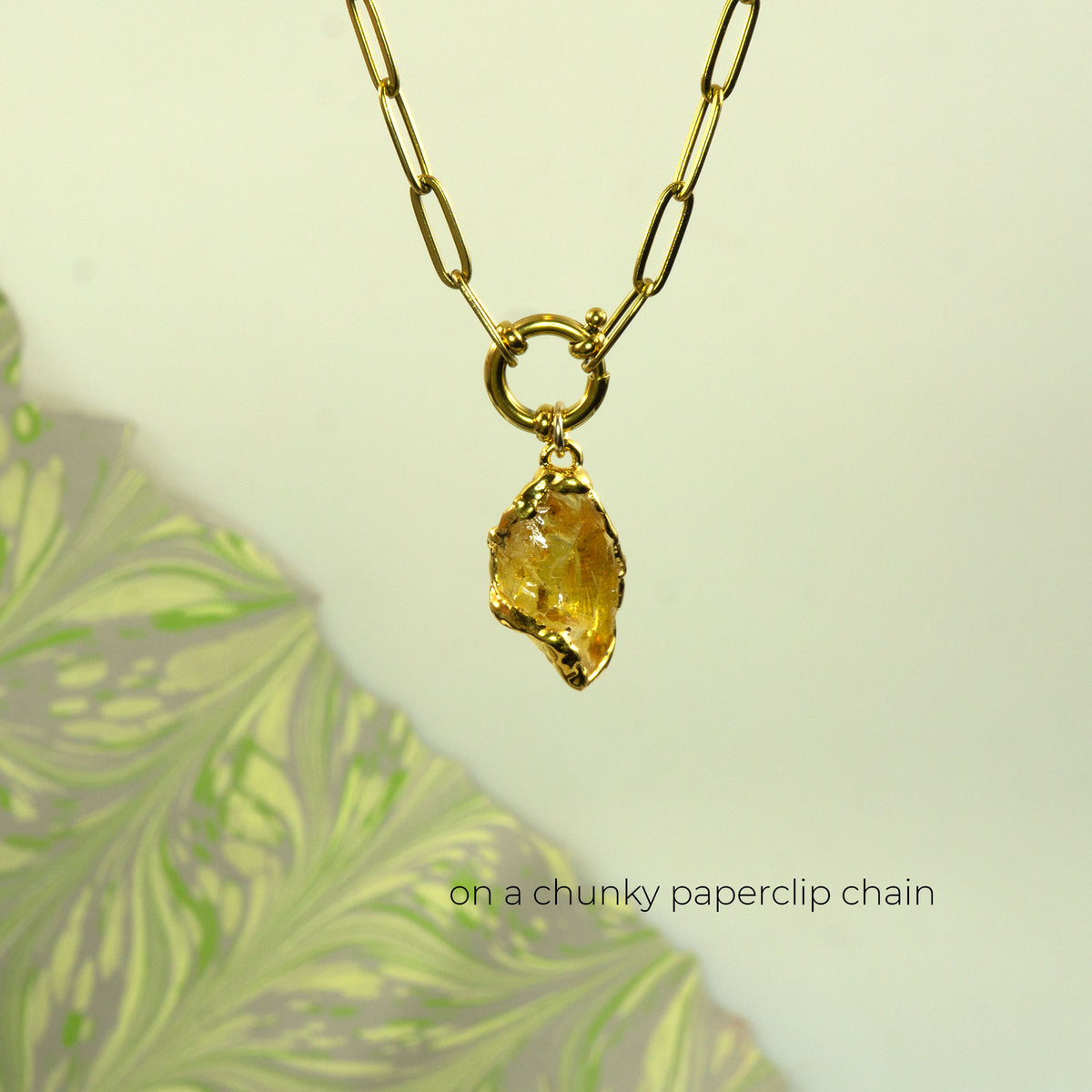A gold necklace with a chunky paperclip chain and a raw, yellow citrine gemstone pendant.