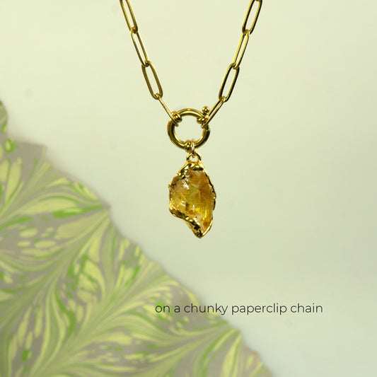 A gold necklace with a chunky paperclip chain and a raw, yellow citrine gemstone pendant.
