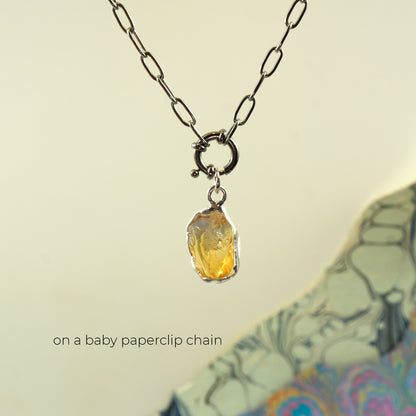 A silver paperclip chain necklace with a small ring clasp holding an irregular, rough-cut yellow citrine gemstone pendant