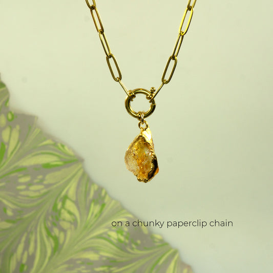 A gold necklace with a raw, irregular yellow citrine gemstone pendant, suspended on a chunky paperclip chain.