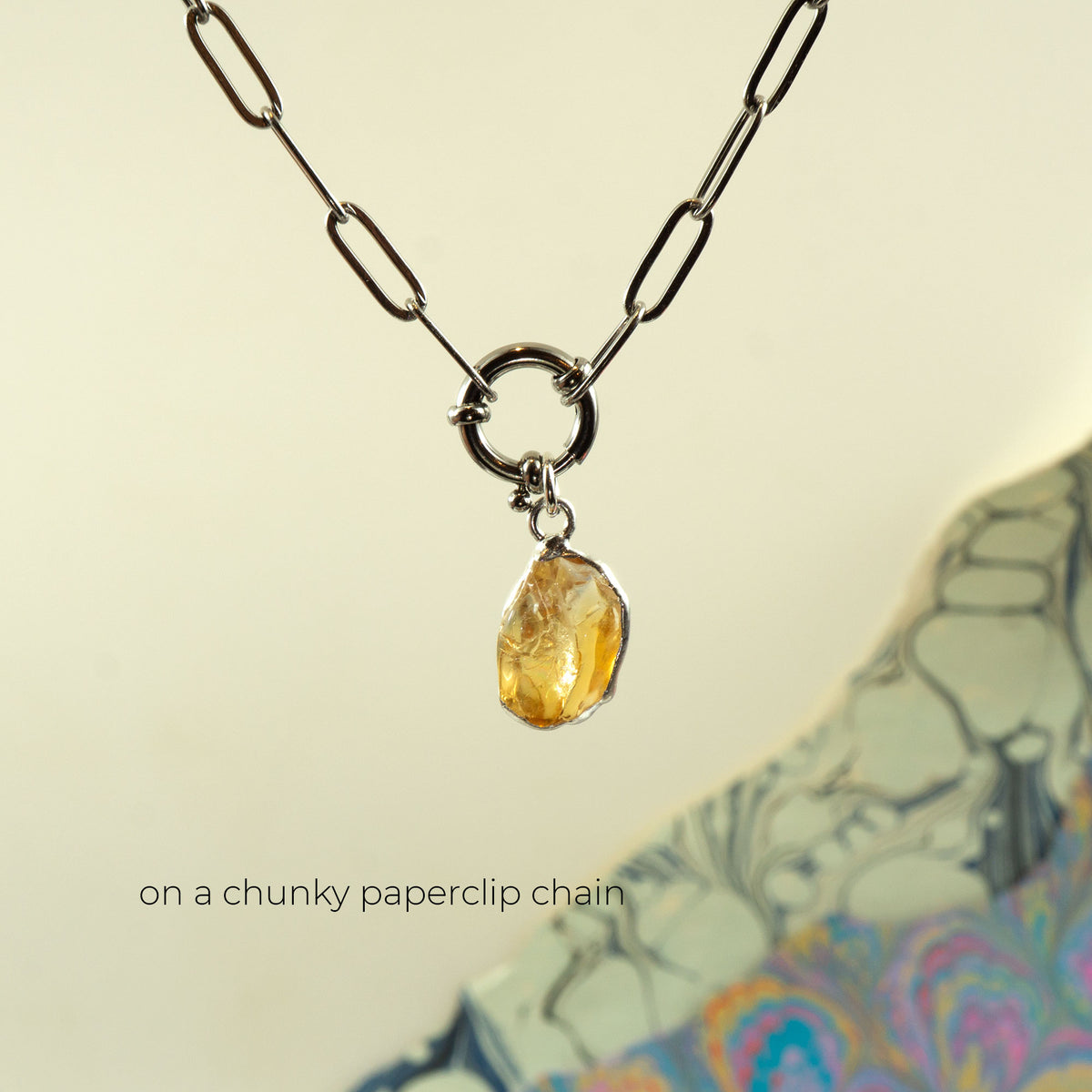 A silver chunky paperclip chain necklace with a ring clasp holding a small, rough, faceted yellow citrine gemstone pendant