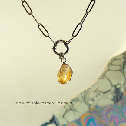 A silver chunky paperclip chain necklace with a ring clasp holding a small, rough, faceted yellow citrine gemstone pendant