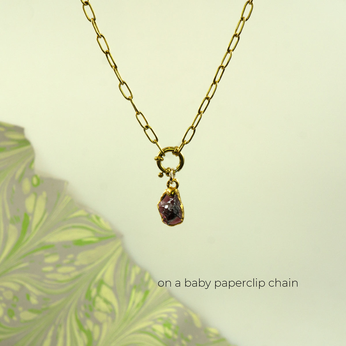 A gold paperclip chain necklace with a small gold ring clasp holding a raw, faceted purple garnet gemstone pendant.