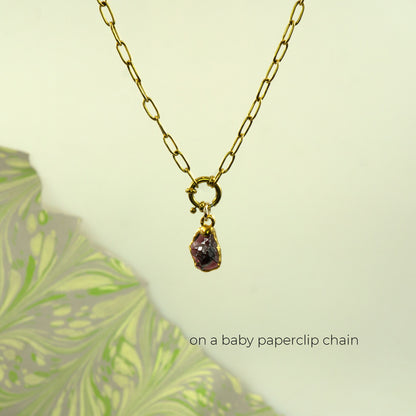 A gold paperclip chain necklace with a small gold ring clasp holding a raw, faceted purple garnet gemstone pendant.