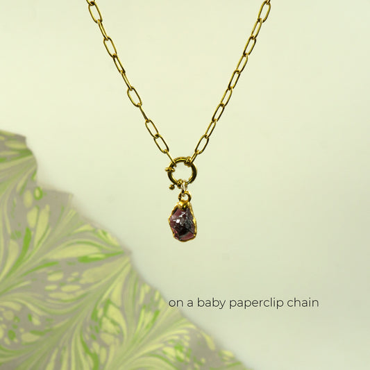 A gold paperclip chain necklace with a small gold ring clasp holding a raw, faceted purple garnet gemstone pendant.