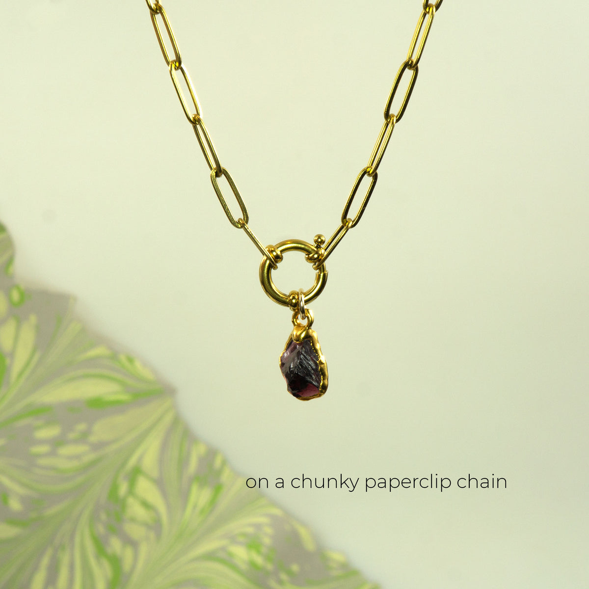 A gold paperclip chain necklace with a small ring connector and a teardrop-shaped dark purple garnet gemstone pendant