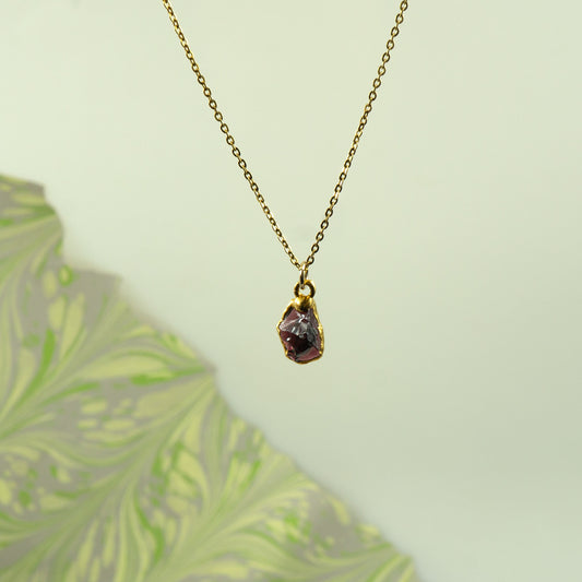 A gold chain necklace with a small, rough-cut dark purple garnet gemstone pendant