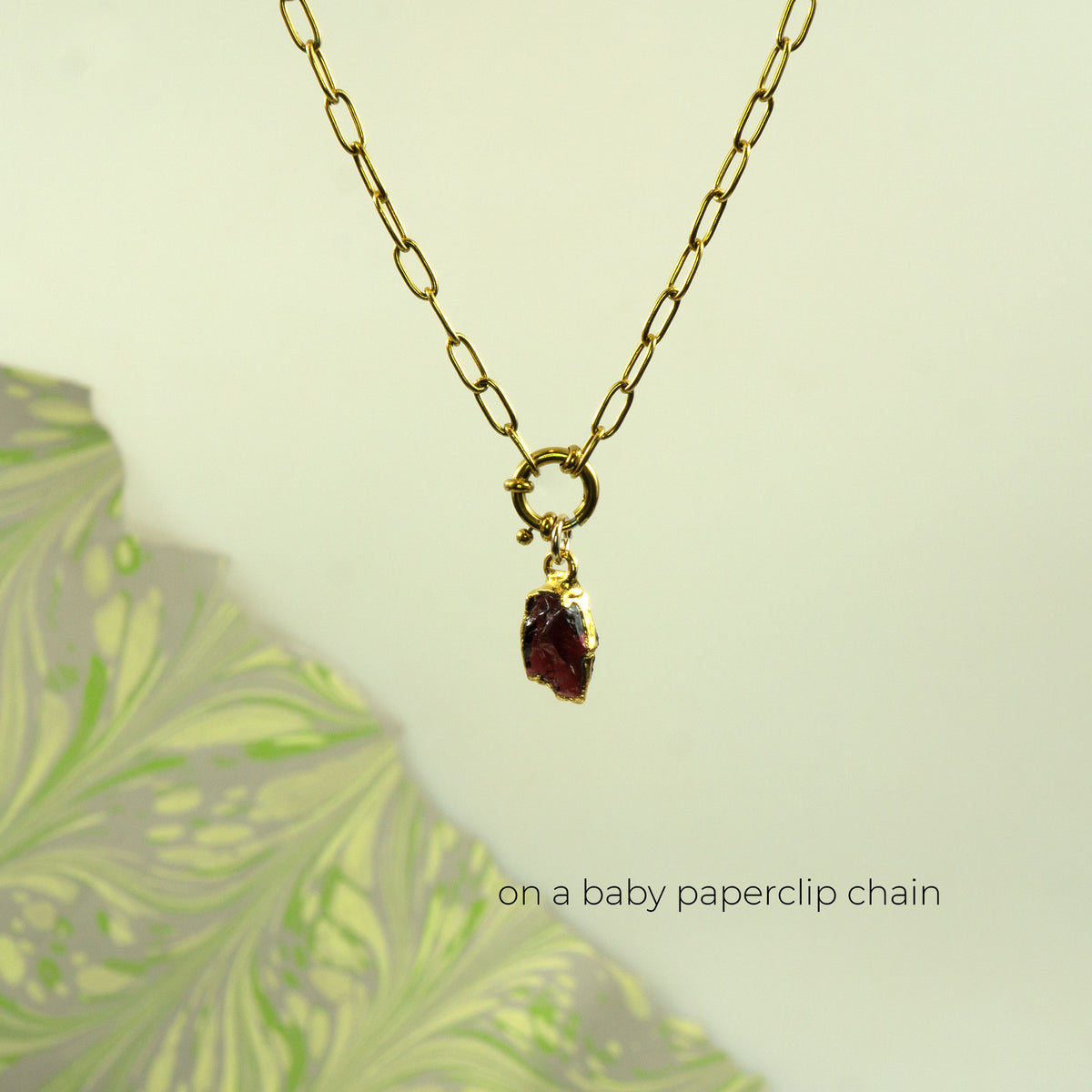A gold paperclip chain necklace with a small bezel-set dark red raw garnet gemstone pendant