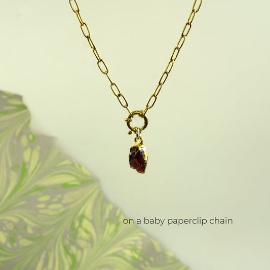 A gold paperclip chain necklace with a small bezel-set dark red raw garnet gemstone pendant