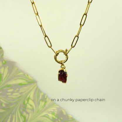 A gold paperclip chain necklace with a small ring clasp holding a rough, dark red garnet gemstone pendant