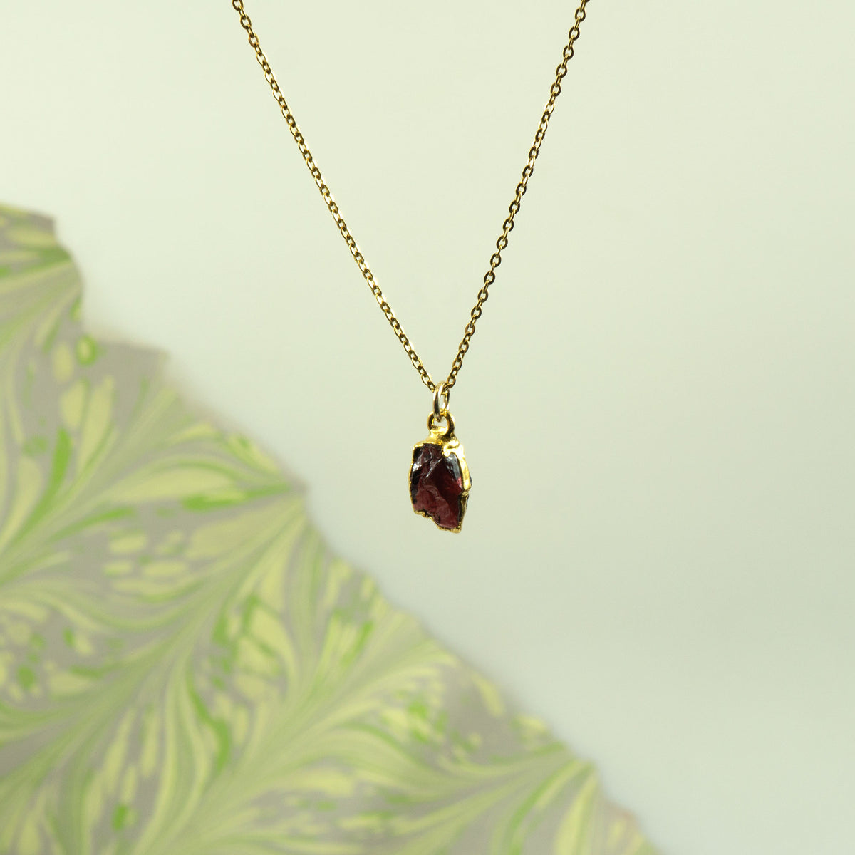 A gold chain necklace with a small, rough-cut dark red garnet gemstone pendant