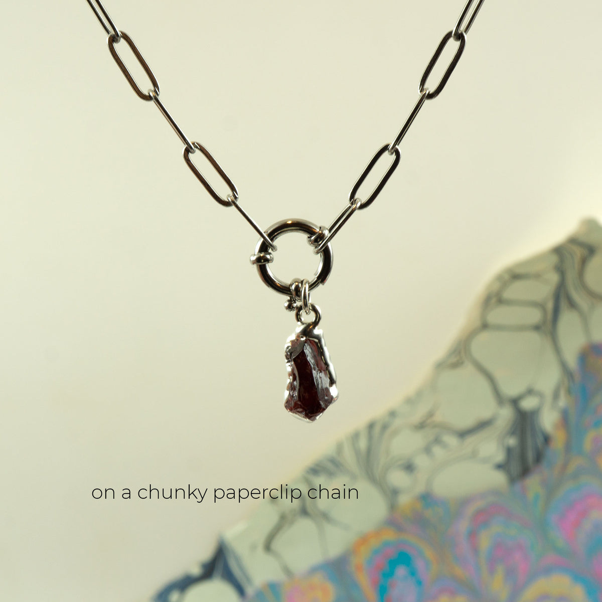 A silver paperclip chain necklace with a small ring clasp holding a dark, faceted crystal garnet pendant