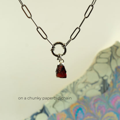 A silver paperclip chain necklace with a circular ring connector and a small raw, faceted dark garnet gemstone pendant