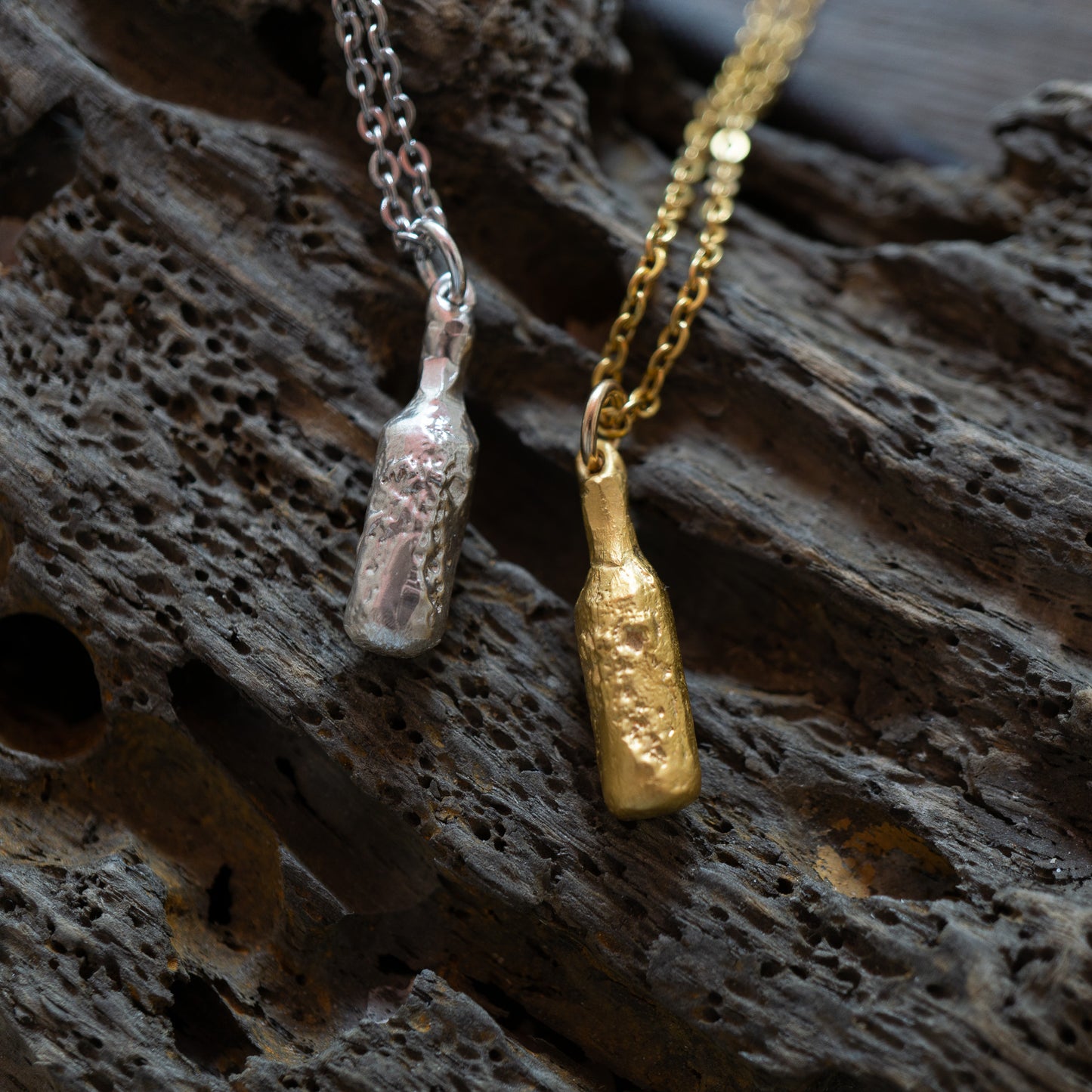 Gold and silver bottle of beer pendants on chains against a textured dark background