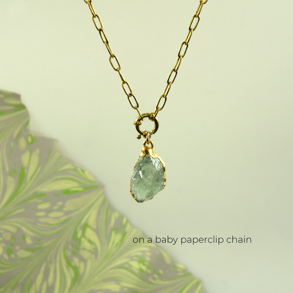 A gold paperclip chain necklace with a small gold clasp holding a raw, faceted Green Amethyst gemstone pendant