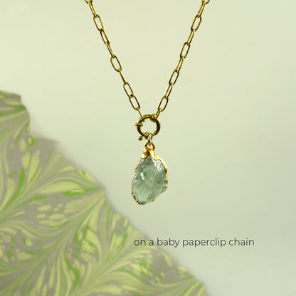 A gold paperclip chain necklace with a small gold clasp holding a raw, faceted Green Amethyst gemstone pendant