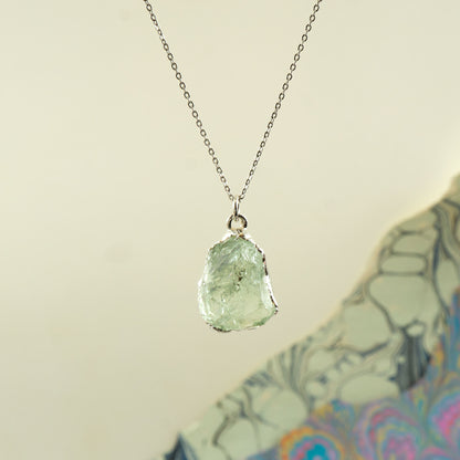 A silver chain necklace with a raw, irregular-cut light Green Amethyst gemstone pendant,
