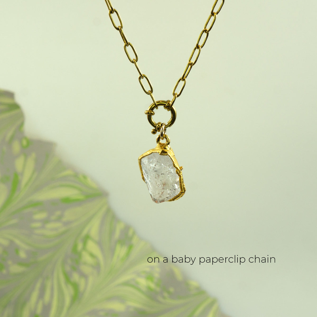 A gold paperclip chain necklace with a raw, rough-cut clear Herkimer Diamond pendant in a gold bezel setting