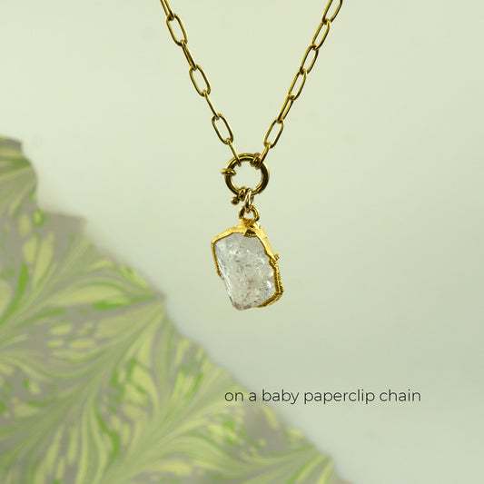 A gold paperclip chain necklace with a raw, rough-cut clear Herkimer Diamond pendant in a gold bezel setting