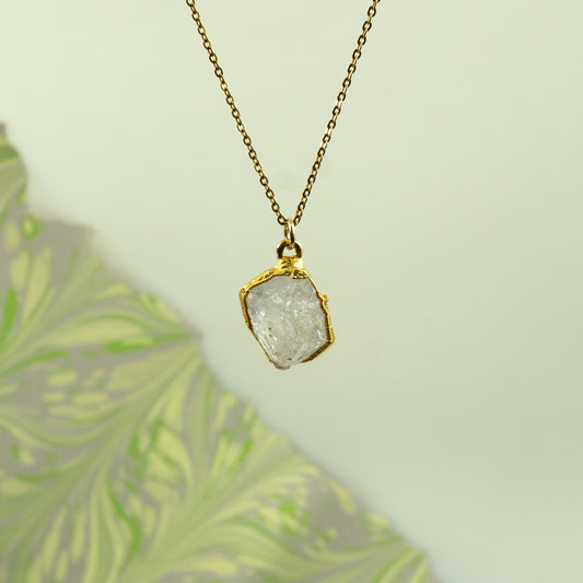 A gold chain necklace with a raw, irregular-cut clear Herkimer Diamond pendant in a gold bezel setting