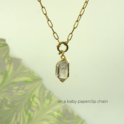 A gold paperclip chain necklace with a small gold clasp holding a clear faceted Herkimer Diamond crystal pendant