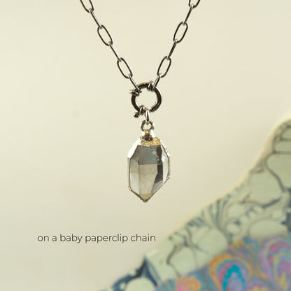 A faceted clear Herkimer diamond crystal pendant necklace on a silver paperclip chain, with a small gold accent at the top of the pendant. 
