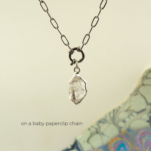 A silver paperclip chain necklace holding a rough, clear faceted Herkimer Diamond pendant.