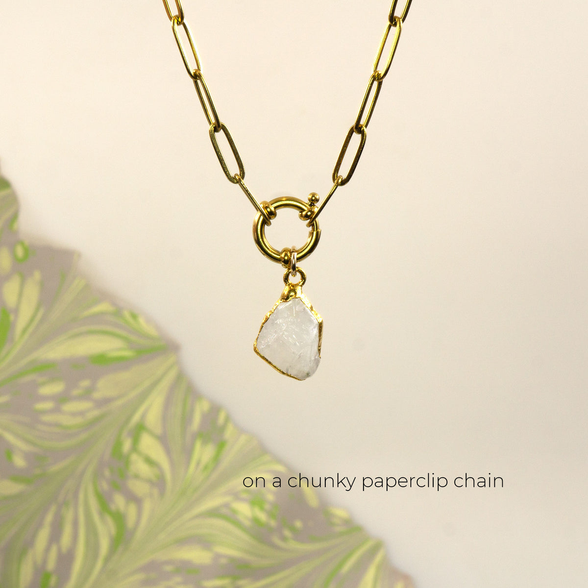 A gold paperclip chain necklace with a gold ring clasp holding a raw, irregular white Rainbow Moonstone gemstone pendant.