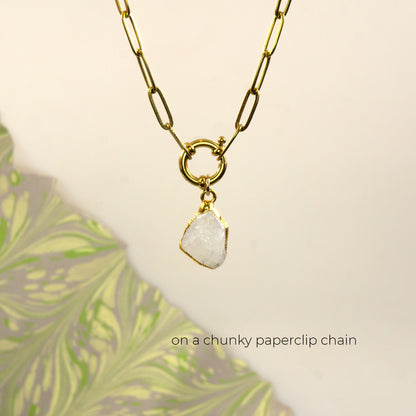 A gold paperclip chain necklace with a gold ring clasp holding a raw, irregular white Rainbow Moonstone gemstone pendant.