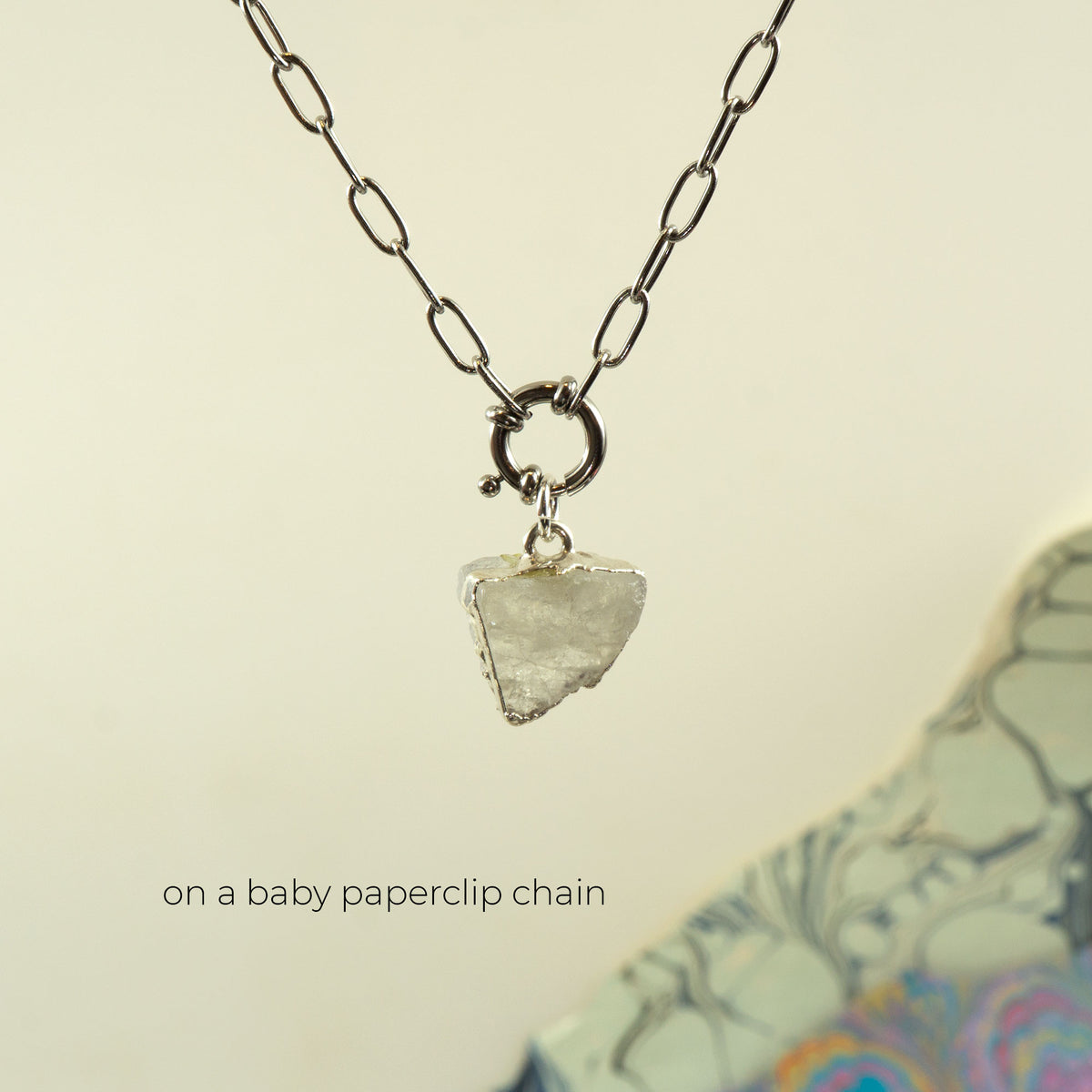 A silver paperclip chain necklace with a small ring clasp holding a rough, irregular clear Rainbow Moonstone pendant