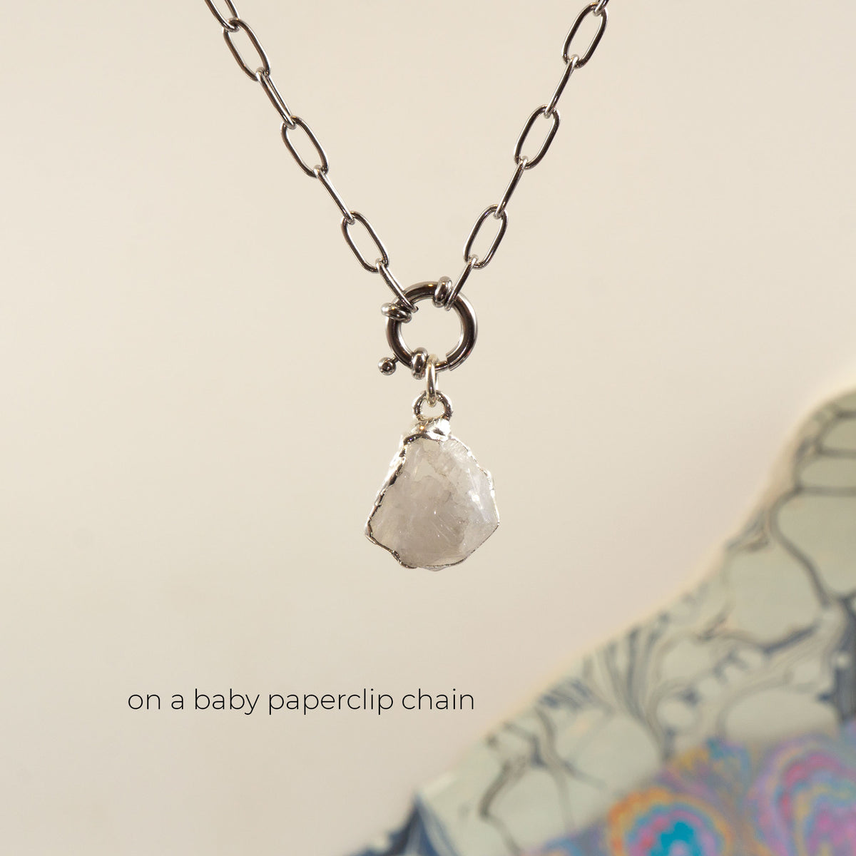 A silver paperclip chain necklace with a large ring connector and a raw, faceted clear Rainbow Moonstone gemstone pendant.