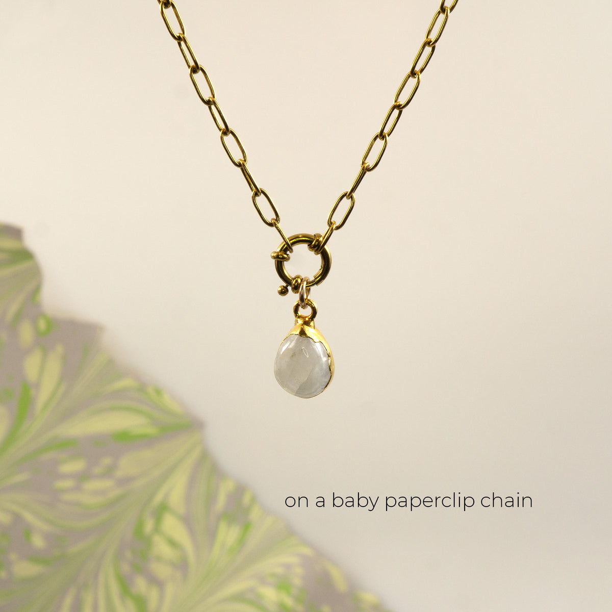 A gold paperclip chain necklace with a small gold ring clasp and a faceted white teardrop Rainbow Moonstone gemstone pendant.