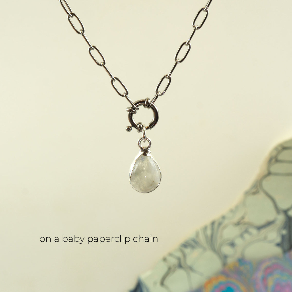 A silver paperclip chain necklace with a small ring clasp and a teardrop-shaped white Rainbow Moonstone pendant