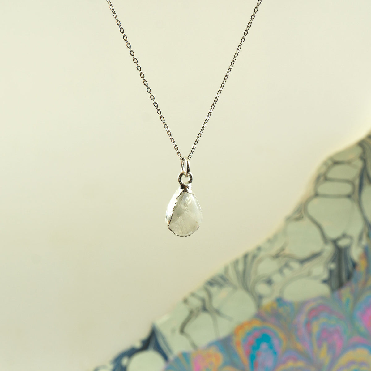 A silver chain necklace with a teardrop-shaped clear faceted Rainbow Moonstone pendant