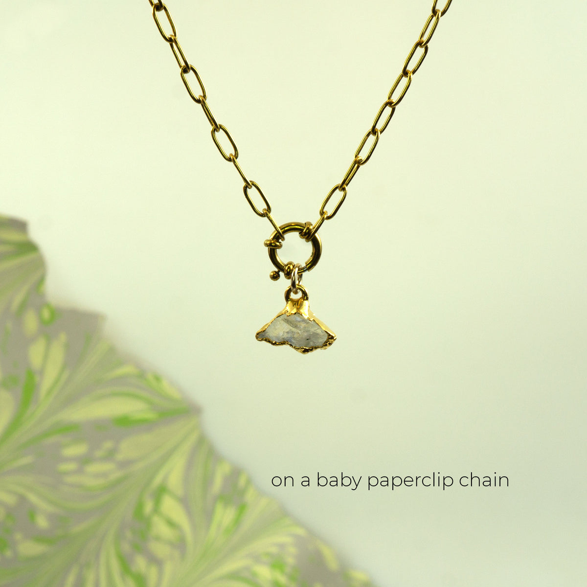 A gold paperclip chain necklace with a small gold ring clasp holding a flat, irregularly shaped pale gray Rainbow Moonstone pendant,.