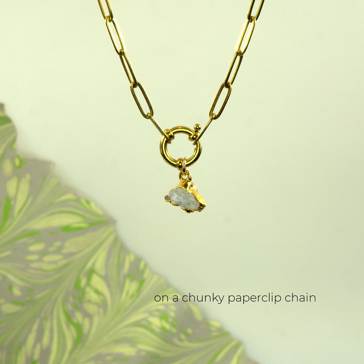 A gold paperclip chain necklace with a ring clasp and a small raw, irregularly shaped white Rainbow Moonstone gemstone pendant.