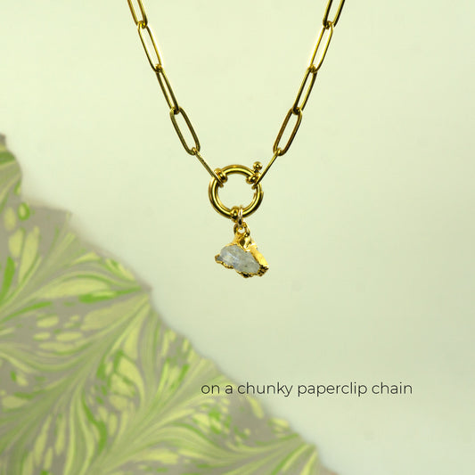 A gold paperclip chain necklace with a ring clasp and a small raw, irregularly shaped white Rainbow Moonstone gemstone pendant.